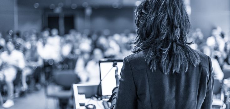 Female speaker giving a talk on corporate business conference. Unrecognizable people in audience at conference hall. Business and Entrepreneurship event. Black and white, blue toned image.