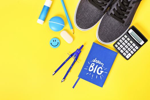 Top view of blue boy match school supplies, sneakers and a "Dream Big" notebook on yellow background.