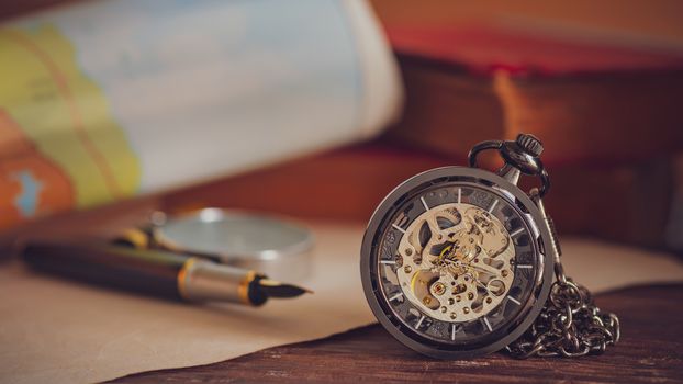 Pocket watch with old books and pen with paper map on the table by the window. Concept of travel planning.