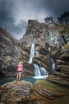 Traveller stands in awe of waterfalls tumbling from over and within imposing rocky limestone gorge and caves flow into stunning blue swimming holes. Moody weather of the Snowy Mountains rumbles overhead