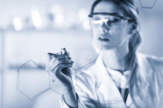 Portrait of a confident female health care expert in life science laboratory writing structural chemical formula on a glass board. Healthcare and modern life science concept. Blue toned image.