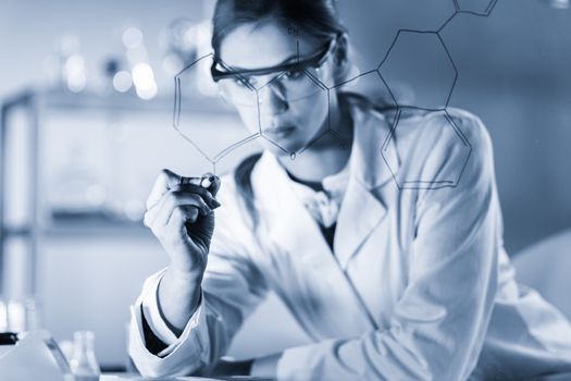 Portrait of a confident female health care expert in life science laboratory writing structural chemical formula on a glass board. Healthcare and modern life science concept. Blue toned image.