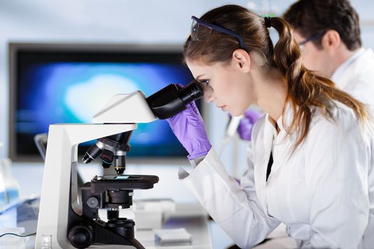 Life scientists researching in laboratory. Attractive female young scientist and her doctoral supervisor microscoping in their working environment. Healthcare and biotechnology.