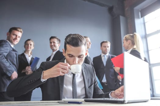 Business man working with laptop and drinkig coffee in office, his team of coworkers on background