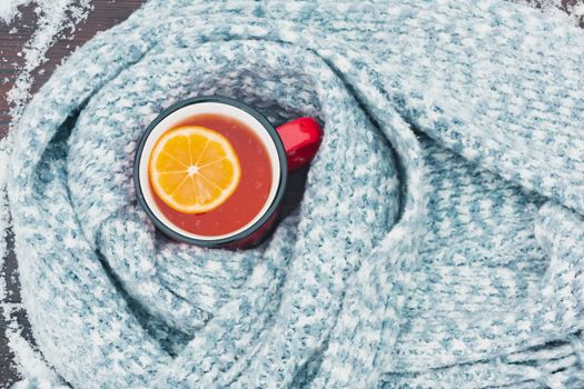 Red enameled cup of hot tea with lemon wrapped in a knitted scarf on a snowy wooden table, top veiw, flat lay.