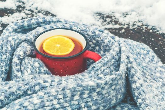 Red enameled cup of hot tea with lemon wrapped in a knitted scarf on a snowy wooden table.