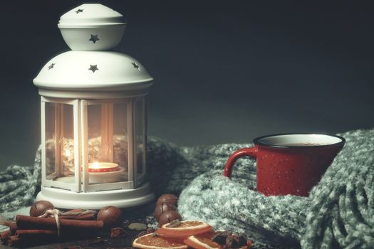 Lantern with a burning candle, spices and a red mug with hot coffee on a snowy wooden table.