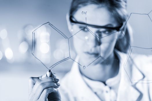 Portrait of a confident female health care expert in life science laboratory writing structural chemical formula on a glass board. Healthcare and modern life science concept. Blue toned image.