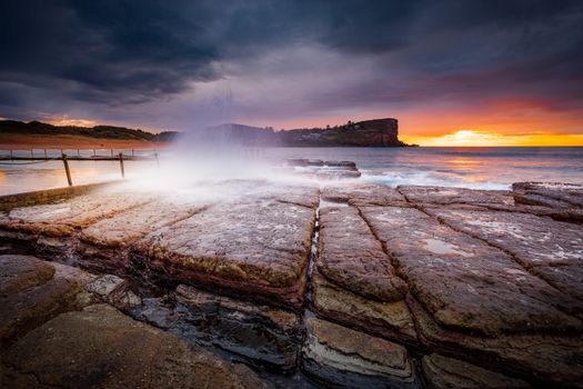 Moody sunrise sky and waves crash onto rocky beach shore.