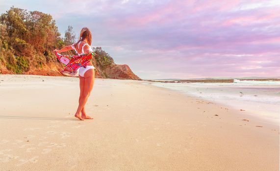 Woman standing on the sandy sea shore with beach towel flapping behind her.  There is some subtle colour in the clouds from the sunrise