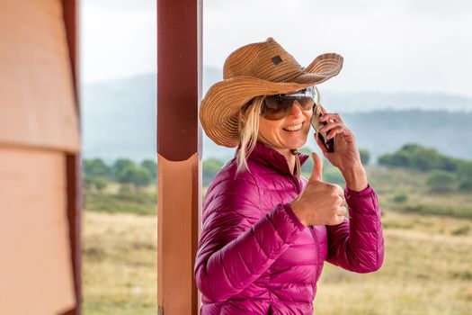 Happy woman standing on porch of rural ranch.  She is using a mobile phone, smiling with thumbs up gesture, RU OK, lifeline, success, etc.