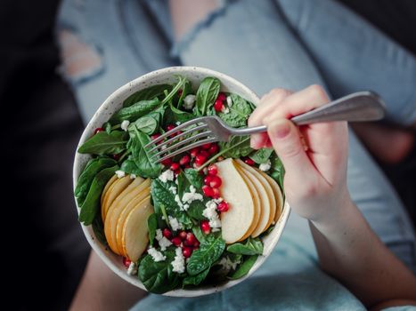 Woman in jeans holding vegan salad bowl with spinach, pear, pomegranate, cheese. Vegan breakfast, vegetarian food, diet concept. Girl in jeans holding fork with knees and hands visible
