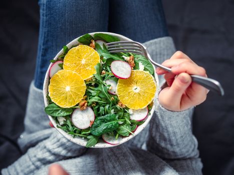 Woman in jeans at bed, holding vegan salad bowl with oranges, spinach,arugula,radish,nut.Top view.Vegan breakfast,vegetarian food,diet concept.Girl in jeans holding fork with knees and hands visible