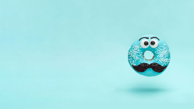 Blue glazed donut with mustache. Flying blue doughnut with funny face with mustache over blue background. Copy space for text. Masculinity or father day concept. Banner