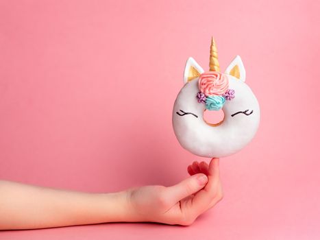 Unicorn donut on forefinger over pink background. Female hand hold trendy donut unicorn with white glaze on index finger. Copy space for text.
