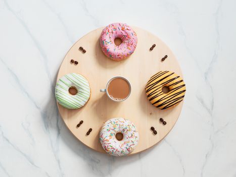 Coffee time and donuts time concept. Clock face with numbers of dougnuts and coffee beans, clock hands as cup of coffee. Wooden tray as watch face, on white marble background. Top view or flat lay.