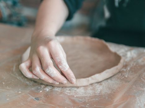 Female ceramist hands make trendy plate of brown clay. Close up view of fingers made fashion dessert flat plate on craft workshop.