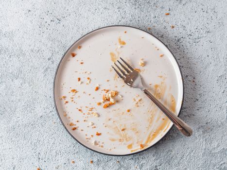 Empty dirty dish after cheesecake with caramel sauce and dessert fork. White rustic trendy modern fashionable dirty plate on gray cement background, top view or flat lay. Copy space for text