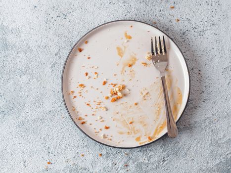 Empty dirty dish after cheesecake with caramel sauce and dessert fork. White rustic trendy modern fashionable dirty plate on gray cement background, top view or flat lay. Copy space for text