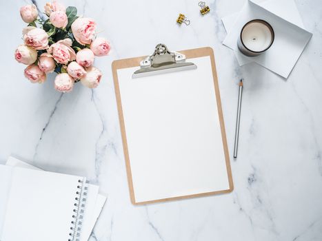 Top view of clipboard with white empty page. Clipboard, flowers, scented candle on white marble. Feminine home office mock up with blank sheet of paper A4 portrait format,copy space for text. Flat lay
