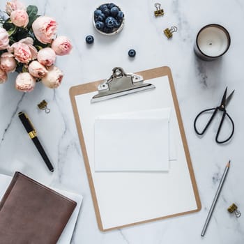 Wedding mock up. Flat lay with blank greeting card, envelope. Clipboard, flowers, scented candle on white marble. Feminine home office mock up with copy space for text or design. Flat lay