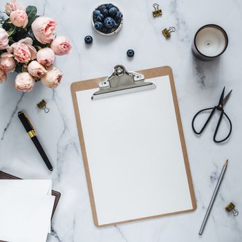 Top view of clipboard with white empty page. Clipboard, flowers, scented candle on white marble. Feminine home office mock up with blank sheet of paper A4 portrait format,copy space for text. Flat lay