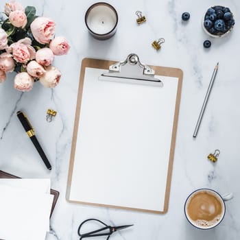 Top view of clipboard with white empty page. Clipboard, flowers, scented candle on white marble. Feminine home office mock up with blank sheet of paper A4 portrait format,copy space for text. Flat lay