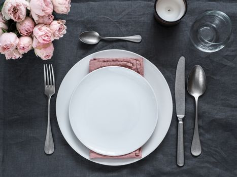 Beautiful table setting on gray linen tablecloth. Festive table setting for wedding dinner with pink spray roses and scented cabdle. Holiday dinner with white plates. Dinner, utensil. Copy space