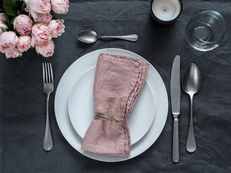 Beautiful table setting with pink spray roses and candle on gray linen tablecloth. Festive table setting for wedding dinner with pink napkin on plate. Holiday dinner with white plates