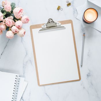 Top view of clipboard with white empty page. Clipboard, flowers, scented candle on white marble. Feminine home office mock up with blank sheet of paper A4 portrait format,copy space for text. Flat lay