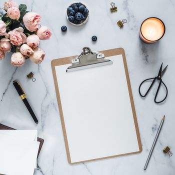 Top view of clipboard with white empty page. Clipboard, flowers, scented candle on white marble. Feminine home office mock up with blank sheet of paper A4 portrait format,copy space for text. Flat lay