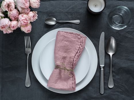 Beautiful table setting with pink spray roses and candle on gray linen tablecloth. Festive table setting for wedding dinner with pink napkin on plate. Holiday dinner with white plates