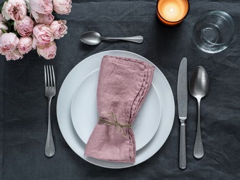 Beautiful table setting with pink spray roses and candle on gray linen tablecloth. Festive table setting for wedding dinner with pink napkin on plate. Holiday dinner with white plates