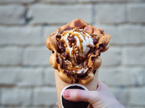 Hong Kong waffles in female hand on brick wall background. Delicious bubble waffle with ice cream and caramel syrup in woman hand. Trendy food.