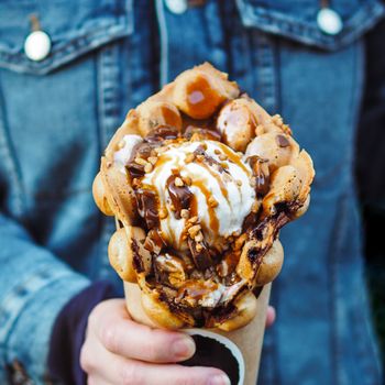 Hong Kong waffles in female hand. Girl in denim jacket hold delicious bubble waffle with ice cream and caramel syrup. Trendy food. Square
