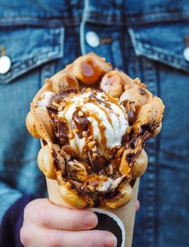 Hong Kong waffles in female hand. Girl in denim jacket hold delicious bubble waffle with ice cream and caramel syrup. Trendy food. Vertical