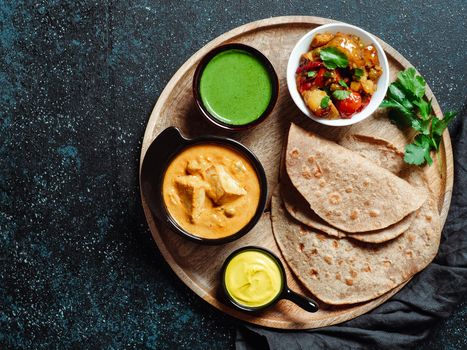 Indian cuisine dishes: vegetable curry, shahi paneer, chapati, chutney. Indian food on wooden tray over dark background. Assortment indian meal with copy space for text. Top view or flat lay.