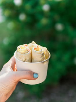 Rolled ice cream in cone cup in woman hand. Woman holds cone cup with thai style kiwi banana rolled ice cream on green leaves background. Outdoor. Natural daylight. Vertical. Copy space for text