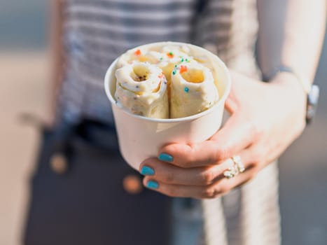 Rolled ice cream in cone cup in woman hands. Woman in striped dress holds cone cup with thai style kiwi banana rolled ice cream.