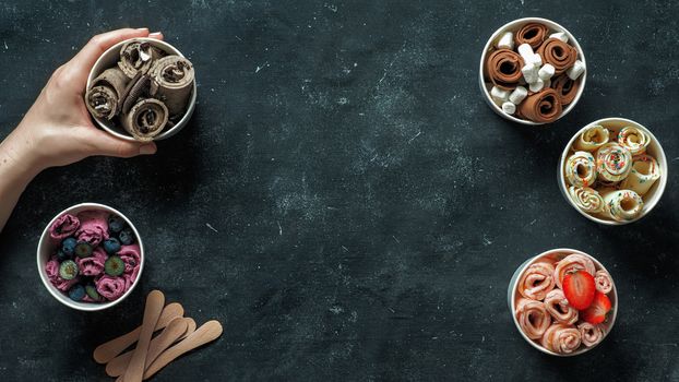 Hand with rolled ice cream in cone cup on dark background. Different iced rolls top view or flat lay. Thai style rolled ice cream with copy space in center for text or design. Banner