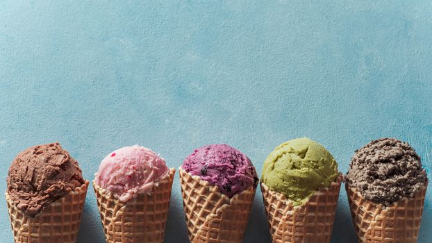 Various ice cream scoops in cones with copy space. Colorful ice cream in cones chocolate, strawberry, blueberry, pistachio or matcha, biscuits chocolate sandwich cookies on blue background. Top view