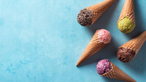 Various ice cream scoops in cones with copy space. Colorful ice cream in cones chocolate, strawberry, blueberry, pistachio or matcha, biscuits chocolate sandwich cookies on blue background. Top view