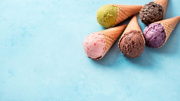 Various ice cream in cones with copy space. Colorful ice cream in cones: chocolate, strawberry, blueberry, pistachio or matcha tea, biscuits chocolate cookies sandwich on blue background. Banner