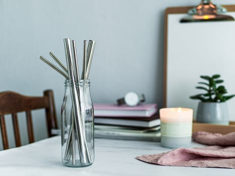 Metal drinking straws in glass bottle on white marble table indoor. Metal straws on table in living room interior. Recyclable straws, zero waste concept.