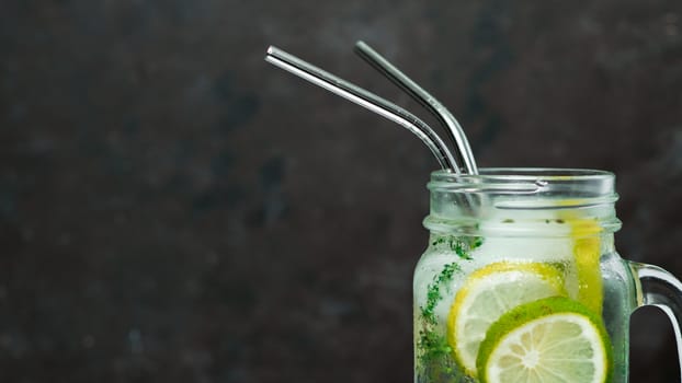 Cold drink in mason jar with metal straw on black background. Lemonade or detox water with lime and thyme in glass jar with copy space for text or design. Recyclable straws, zero waste concept. Banner