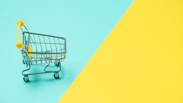 Empty miniature shopping cart on blue and yellow background. Toy trolley on bright colorful background, copy space for text or design.