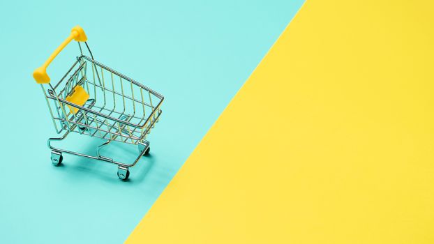 Empty miniature shopping cart on blue and yellow background. Toy trolley on bright colorful background, copy space for text or design.