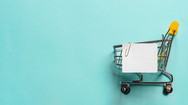 Shopping cart and white paper note list over blue background. Shopping concept on blue background. Empty white paper note over shopping cart. Copy space for text or design. Banner