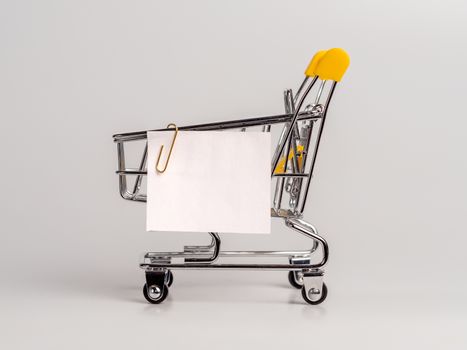 Shopping cart and white paper note list isolated on white background. Shopping concept on light monochrome background. Empty white paper note over shopping cart. Copy space for text or design.