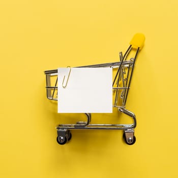 Shopping cart and white paper note list over yellow background. Shopping concept on bright yellow background. Empty white paper note over shopping cart. Copy space for text or design.